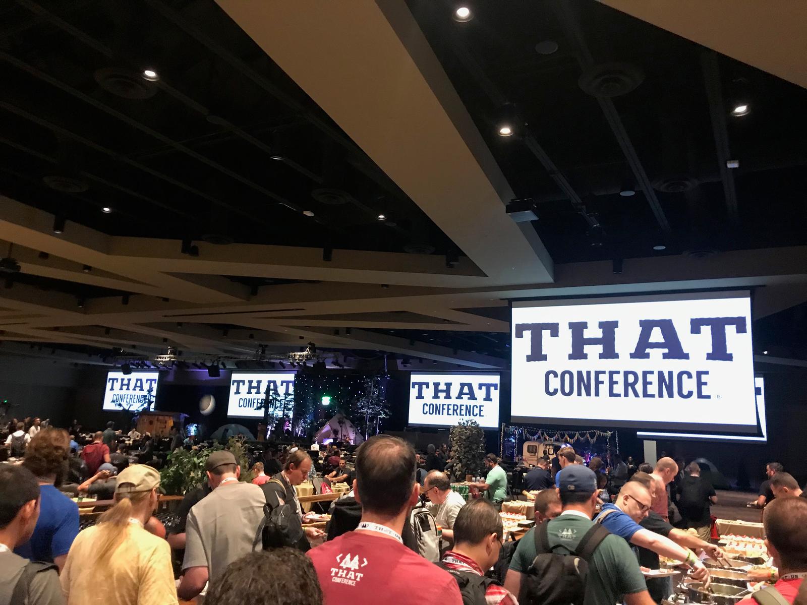 Several attendees stand in line for a meal at THAT conference, the name of the conference is prominently displayed on multiple projectors in the distance.