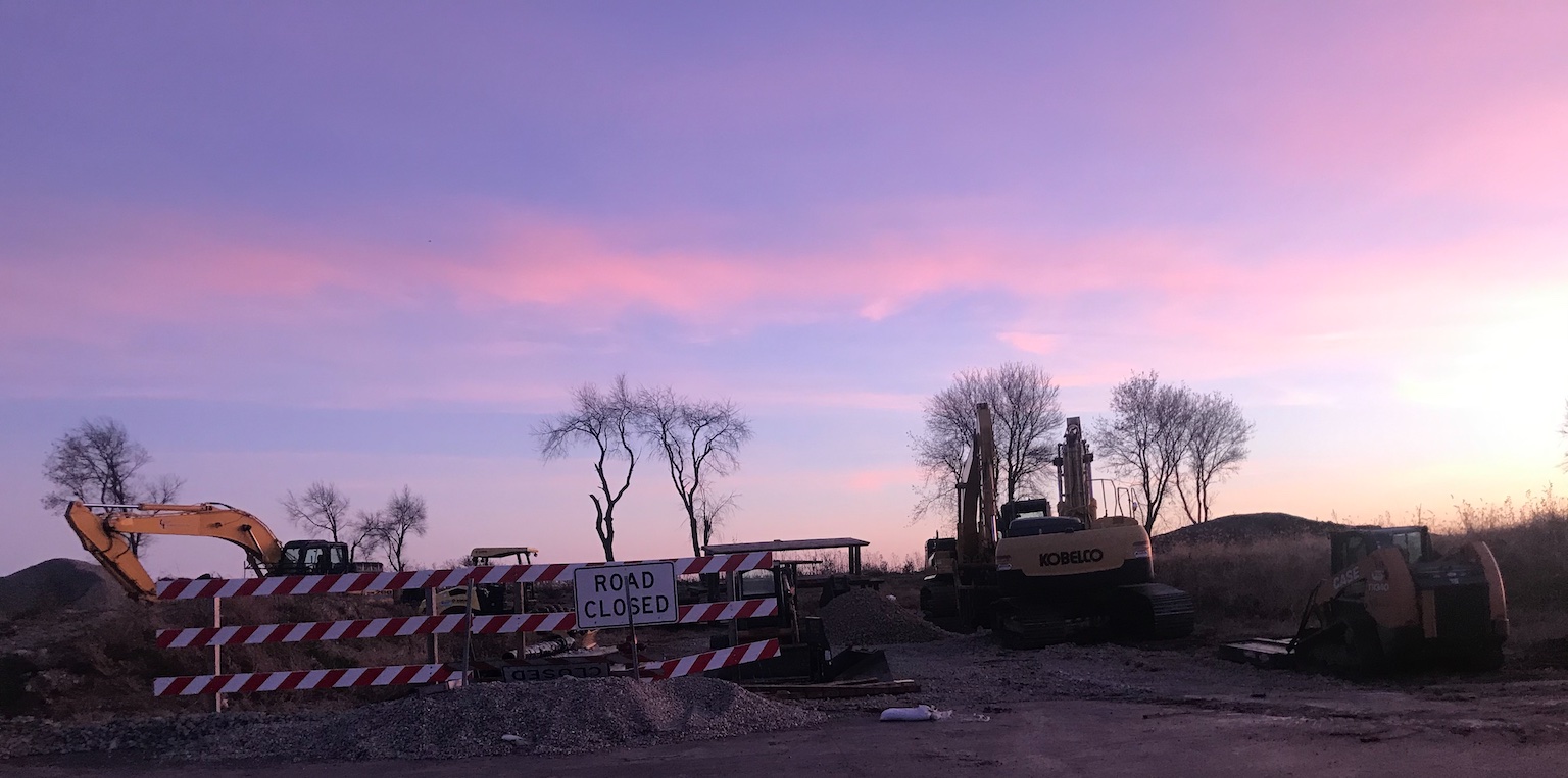 A sign reading road closed next to construction equipment with a very pink sunset behind