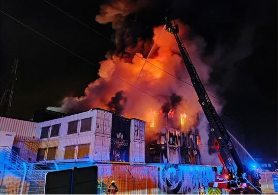 A large fire rages at night within a structure composed of stacked shipping containers, sending thick plumes of smoke and bright orange flames into the sky. A crane arm extends over the burning building as firefighters spray water on the blaze, which is illuminated by the fire's intense glow and blue emergency vehicle lights reflecting off the side of the containers.